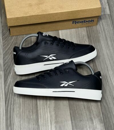 A pair of black and white Reebok Classic Leather sneakers with box on a wooden floor, perfect for men's casual wear.