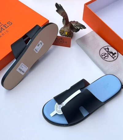 A pair of black leather Hermes Paris slide sandals with light blue insoles, accompanied by an orange box, dust bag, and eagle figurine on a white background.