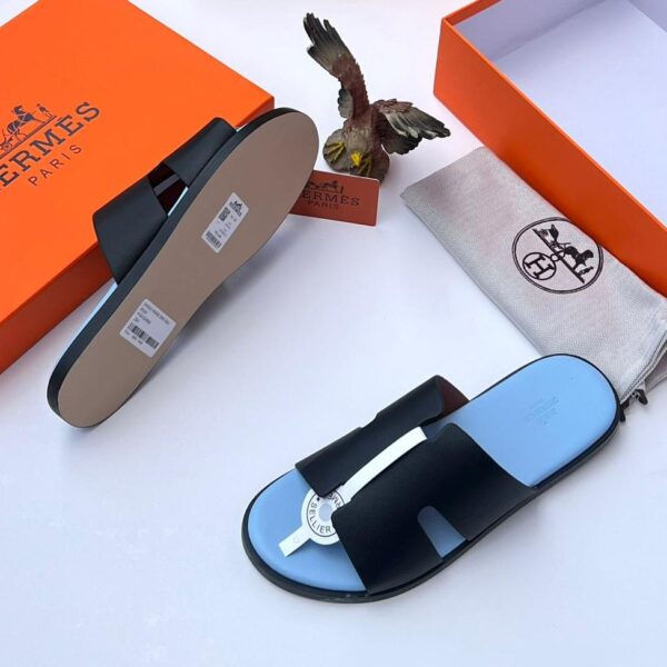 A pair of black leather Hermes Paris slide sandals with light blue insoles, accompanied by an orange box, dust bag, and eagle figurine on a white background.