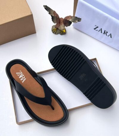 A pair of black T-strap flip flops with brown insole, placed on a white background with a shoe box and bag.