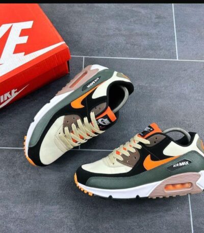 A pair of black, green, and orange Nike Air Max sneakers on a grey tiled floor next to a red Nike shoe box, showcasing the shoe's sleek design and vibrant colors.