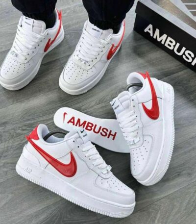A pair of white and red Nike Air Force 1 sneakers with AMBUSH branding, displayed on a grey wooden floor with a black shoe box in the background.