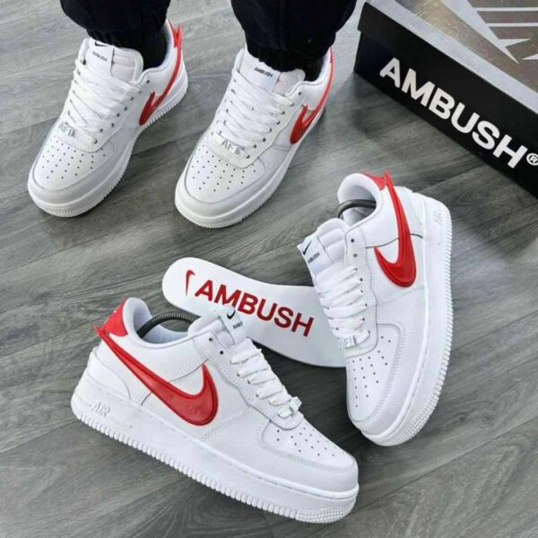 A pair of white and red Nike Air Force 1 sneakers with AMBUSH branding, displayed on a grey wooden floor with a black shoe box in the background.