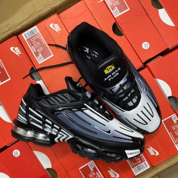 A pair of black and white Nike Air Max Plus sneakers on top of red shoe boxes, showcasing the shoe's sleek design and Tuned Air technology for ultimate comfort and style.