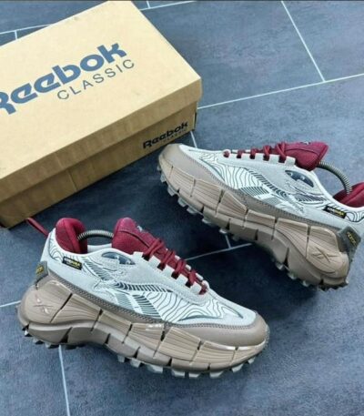A pair of white Reebok Classic sneakers with red accents and aggressive tread pattern, next to a Reebok shoe box on a blue tile floor.