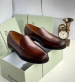 A pair of rich brown leather loafers on a shoe box with a clock in the background, showcasing stylish and comfortable footwear for men.