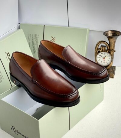 A pair of rich brown leather loafers on a shoe box with a clock in the background, showcasing stylish and comfortable footwear for men.