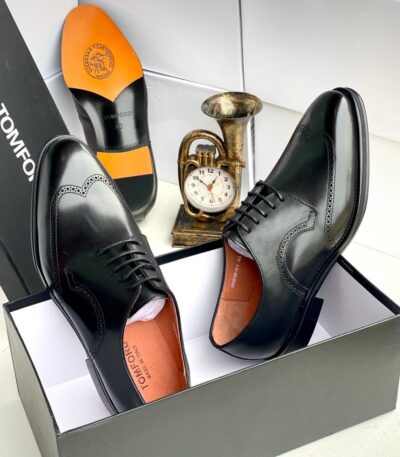 A pair of black leather brogue Oxfords with orange insoles, displayed in a box with a clock and trumpet in the background.