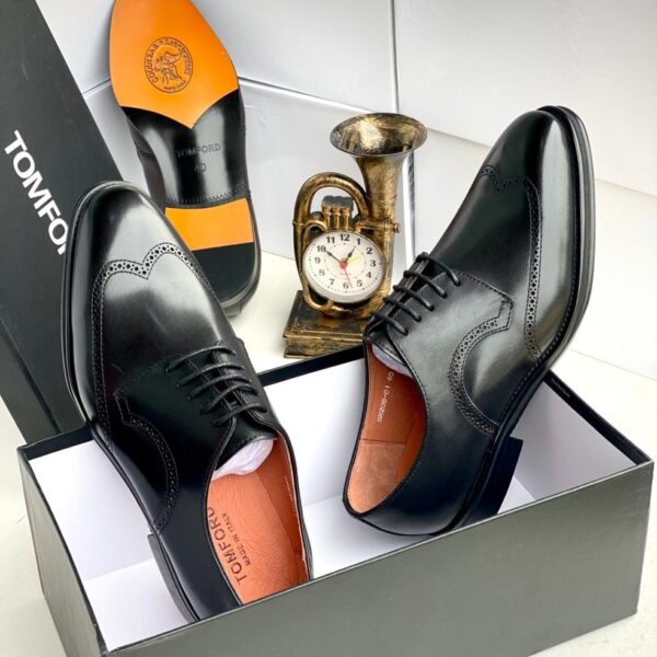 A pair of black leather brogue Oxfords with orange insoles, displayed in a box with a clock and trumpet in the background.