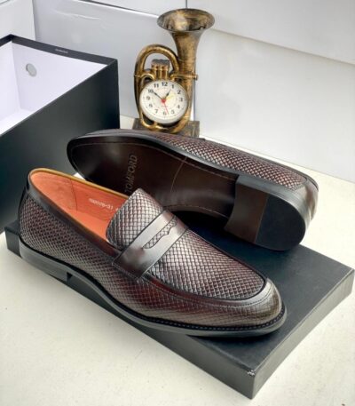 A pair of Tom Ford dark brown leather loafers with intricate texture, displayed on a black shoe box with a gold clock in the background.