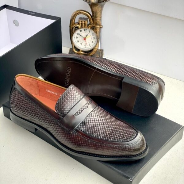 A pair of Tom Ford dark brown leather loafers with intricate texture, displayed on a black shoe box with a gold clock in the background.