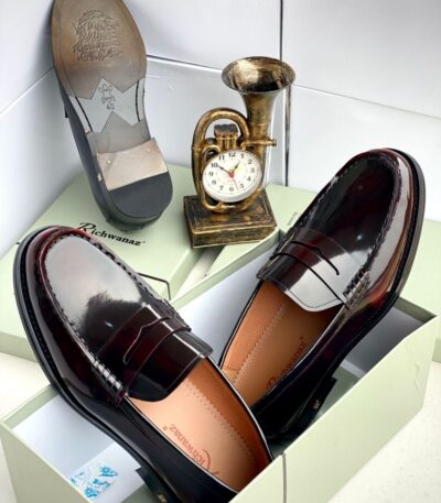 A pair of dark brown leather loafers from Richwanaz, displayed in an open box with a clock and other accessories.