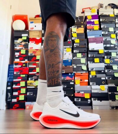 A stylish pair of white Nike running shoes with a black swoosh and red sole, showcased on a man's foot with a unique tattoo, set against a backdrop of shoe boxes.