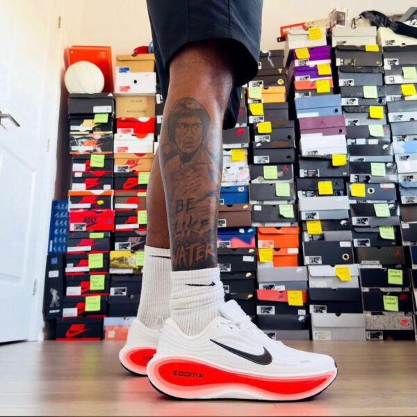 A stylish pair of white Nike running shoes with a black swoosh and red sole, showcased on a man's foot with a unique tattoo, set against a backdrop of shoe boxes.