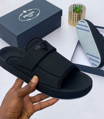 A hand holding a black slide sandal with a triangle logo strap, alongside a shoe box and the sandal's sole, showcasing the luxurious design and attention to detail.