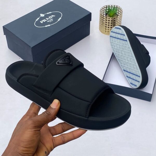 A hand holding a black slide sandal with a triangle logo strap, alongside a shoe box and the sandal's sole, showcasing the luxurious design and attention to detail.