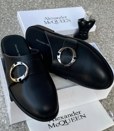 A pair of black leather mule shoes with a gold buckle, placed on top of a white shoe box with the brand name Alexander McQUEEN