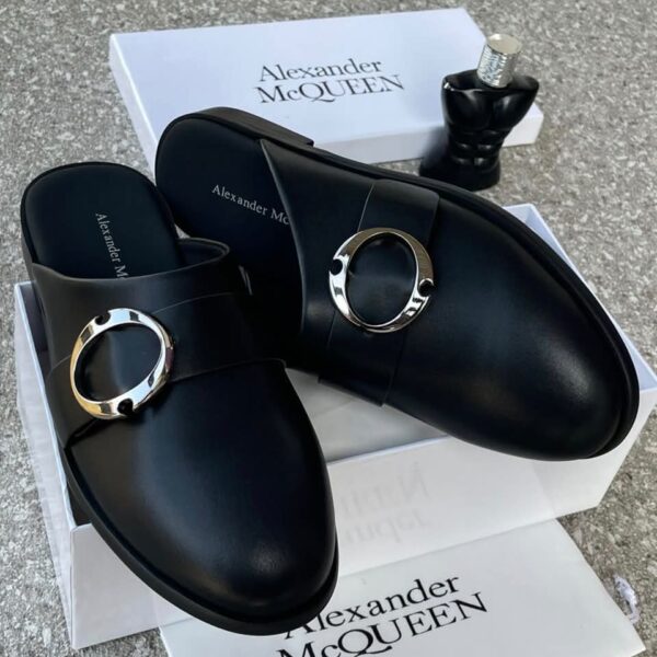 A pair of black leather mule shoes with a gold buckle, placed on top of a white shoe box with the brand name Alexander McQUEEN