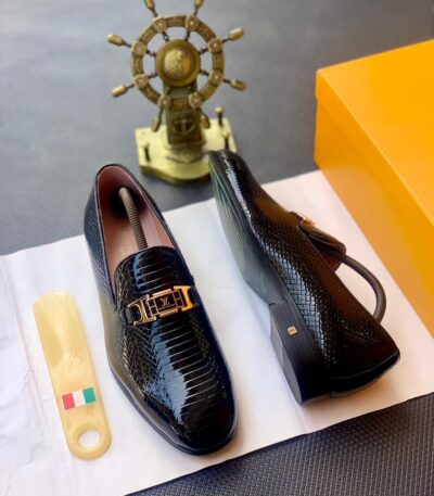 A pair of black snakeskin leather loafers with a gold buckle on a white paper, accompanied by a shoe horn and a golden ship's wheel in the background.