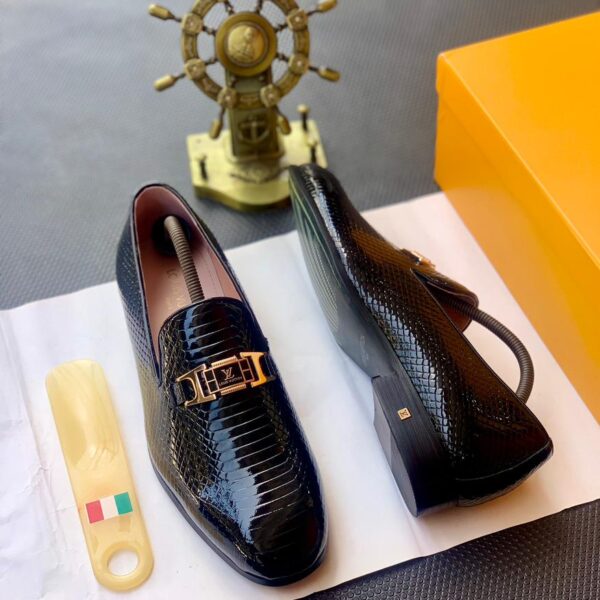 A pair of black snakeskin leather loafers with a gold buckle on a white paper, accompanied by a shoe horn and a golden ship's wheel in the background.