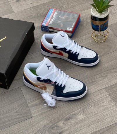 A pair of white, black, and gold Air Jordan 1 Mid SE sneakers on a wooden floor, with a shoe box and gaming console in the background.