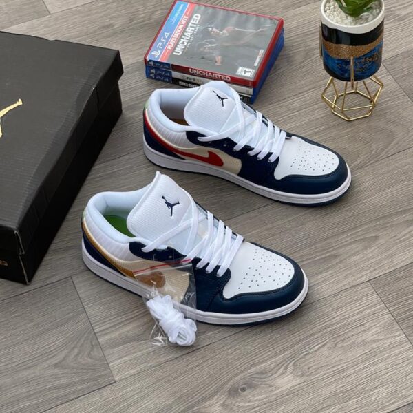 A pair of white, black, and gold Air Jordan 1 Mid SE sneakers on a wooden floor, with a shoe box and gaming console in the background.