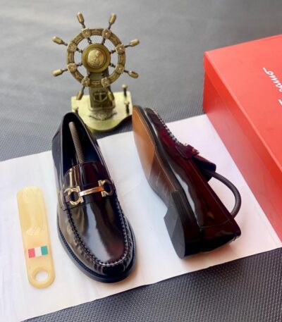 A pair of black leather loafers with a gold buckle on a white paper, accompanied by a shoe care tool and a red shoe box, exuding luxury and sophistication.