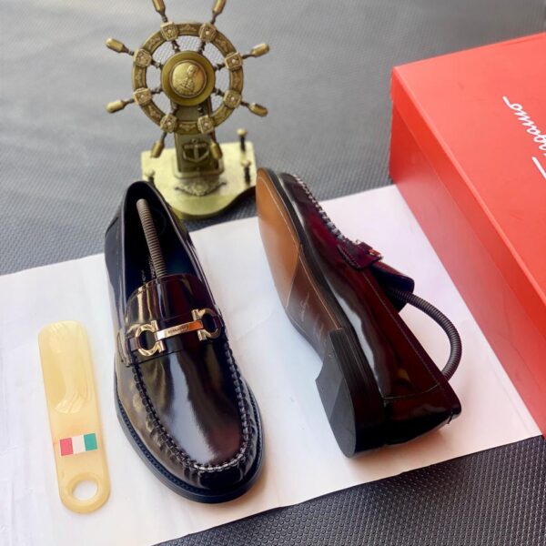 A pair of black leather loafers with a gold buckle on a white paper, accompanied by a shoe care tool and a red shoe box, exuding luxury and sophistication.