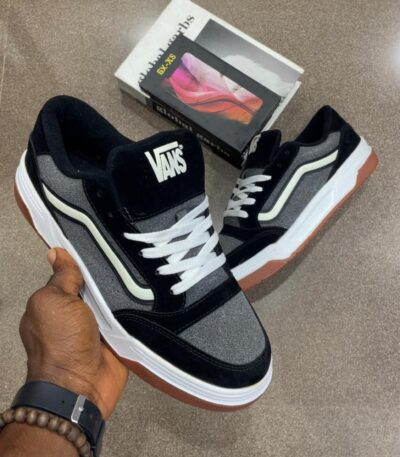 A pair of black and grey Vans sneakers with white laces and brown soles, held by a hand with a watch and bracelet.
