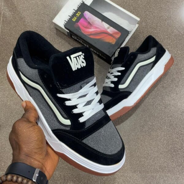 A pair of black and grey Vans sneakers with white laces and brown soles, held by a hand with a watch and bracelet.