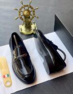 A pair of black patent leather loafers with gold-tone hardware, presented on a white paper with a shoe tree and a black box in the background, embodying the essence of luxury footwear.