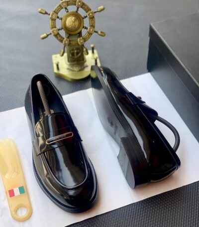 A pair of black patent leather loafers with gold-tone hardware, presented on a white paper with a shoe tree and a black box in the background, embodying the essence of luxury footwear.