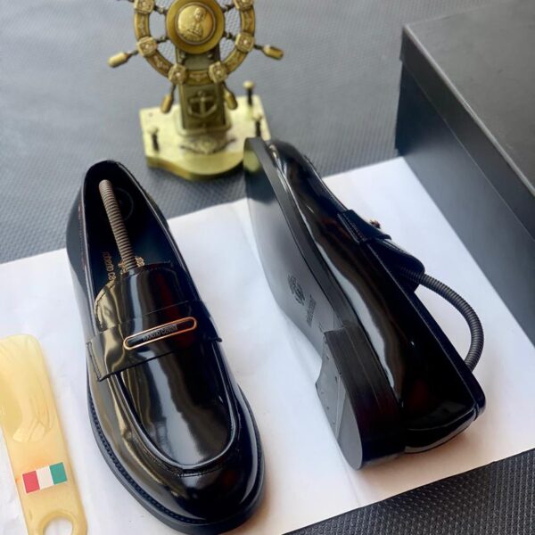 A pair of black patent leather loafers with gold-tone hardware, presented on a white paper with a shoe tree and a black box in the background, embodying the essence of luxury footwear.