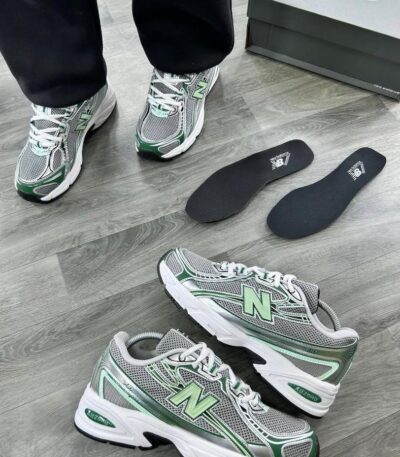 A pair of grey and green New Balance 574 sneakers on a wooden floor, with a shoe box and insoles nearby, showcasing the shoe's sleek design and premium features.