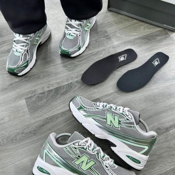 A pair of grey and green New Balance 574 sneakers on a wooden floor, with a shoe box and insoles nearby, showcasing the shoe's sleek design and premium features.