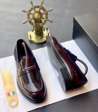 A pair of black patent leather loafers with gold-tone hardware, presented on a white paper with a shoe tree and a black box in the background, embodying the essence of luxury footwear.