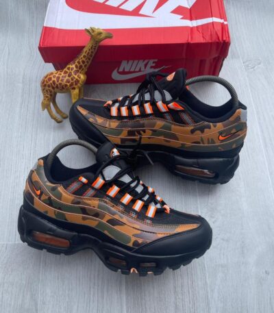 A pair of black and orange camo-print Nike Air Max 270 React sneakers on a grey wooden floor next to a red Nike shoe box and a small giraffe toy.