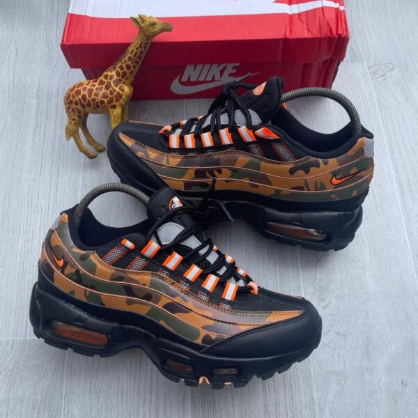 A pair of black and orange camo-print Nike Air Max 270 React sneakers on a grey wooden floor next to a red Nike shoe box and a small giraffe toy.