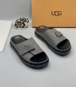 Grey UGG slide sandals with gold accents and stitched sole, presented with a UGG box and luxury accessories.