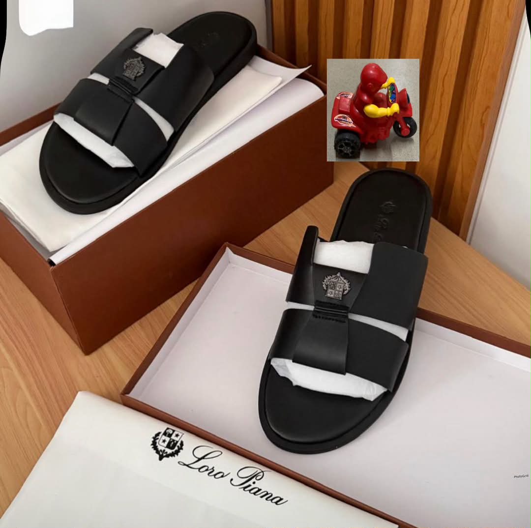 root A pair of luxurious black double-strap slide sandals with embroidered logo, presented on a wooden table with a brown shoe box and white tissue paper, embodying comfort and class.