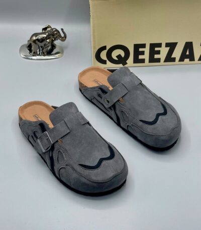 A pair of gray suede double-buckle moccasin shoes on a white background, with a cardboard box and a silver elephant statue in the background.