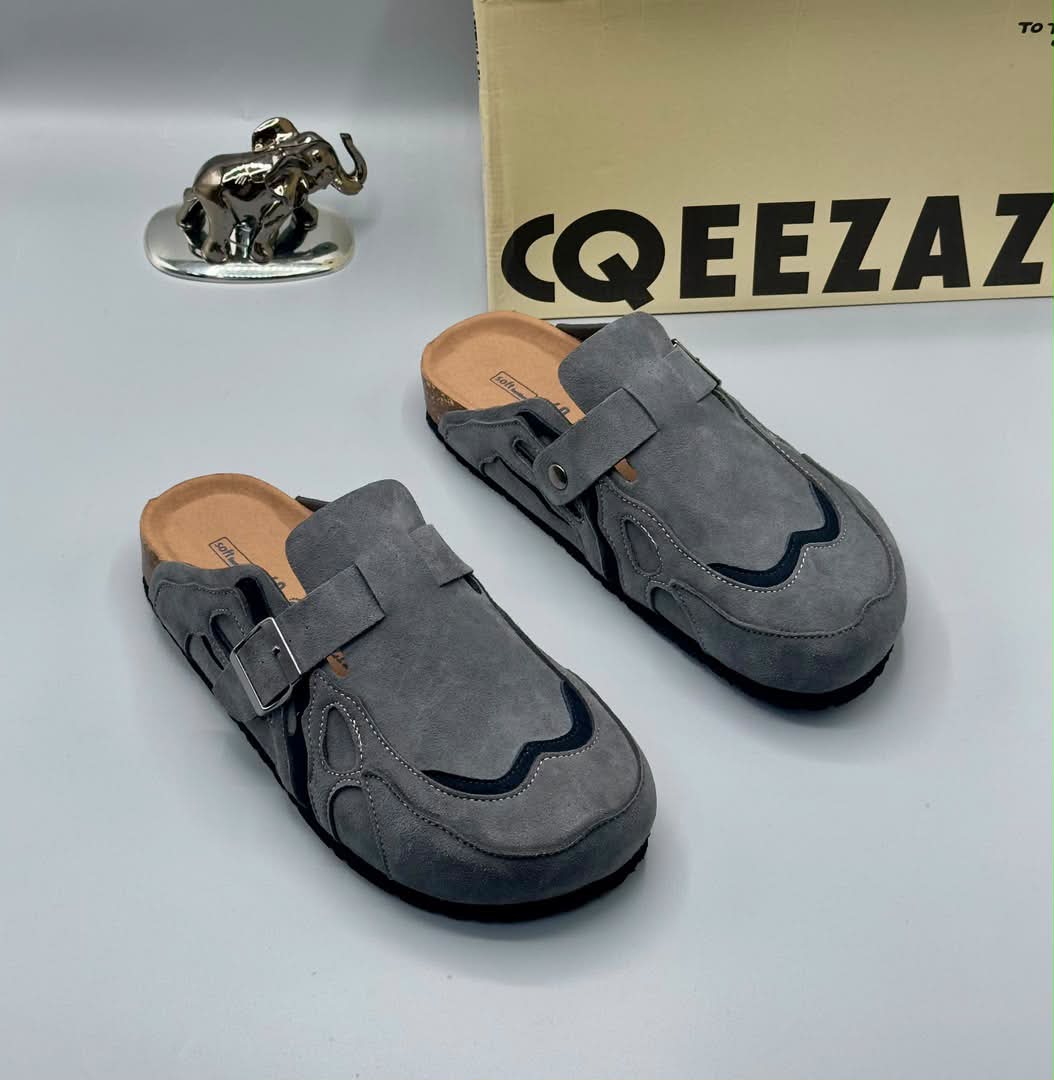 root A pair of gray suede double-buckle moccasin shoes on a white background, with a cardboard box and a silver elephant statue in the background.