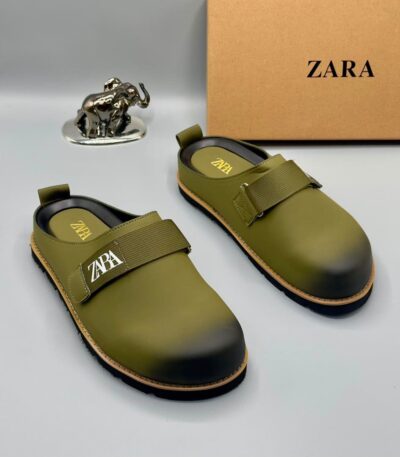 A pair of olive green strap shoes with golden accents, displayed with a silver elephant statue and a Zara box on a white background.