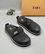 A pair of black leather clogs with gold accents and a velcro strap, displayed on a white surface with a brown box and silver elephant figurine in the background.