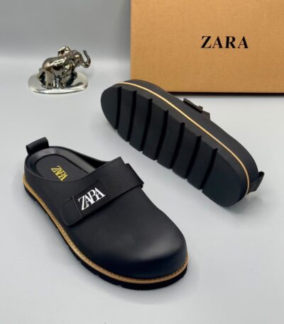 A pair of black leather clogs with gold accents and a velcro strap, displayed on a white surface with a brown box and silver elephant figurine in the background.