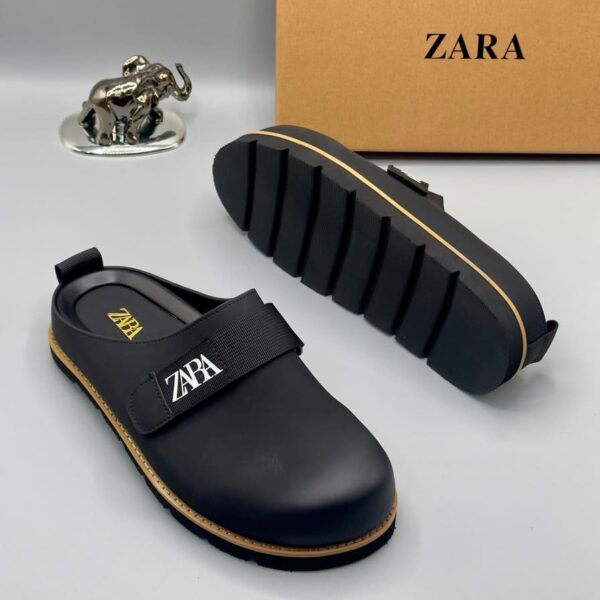 A pair of black leather clogs with gold accents and a velcro strap, displayed on a white surface with a brown box and silver elephant figurine in the background.