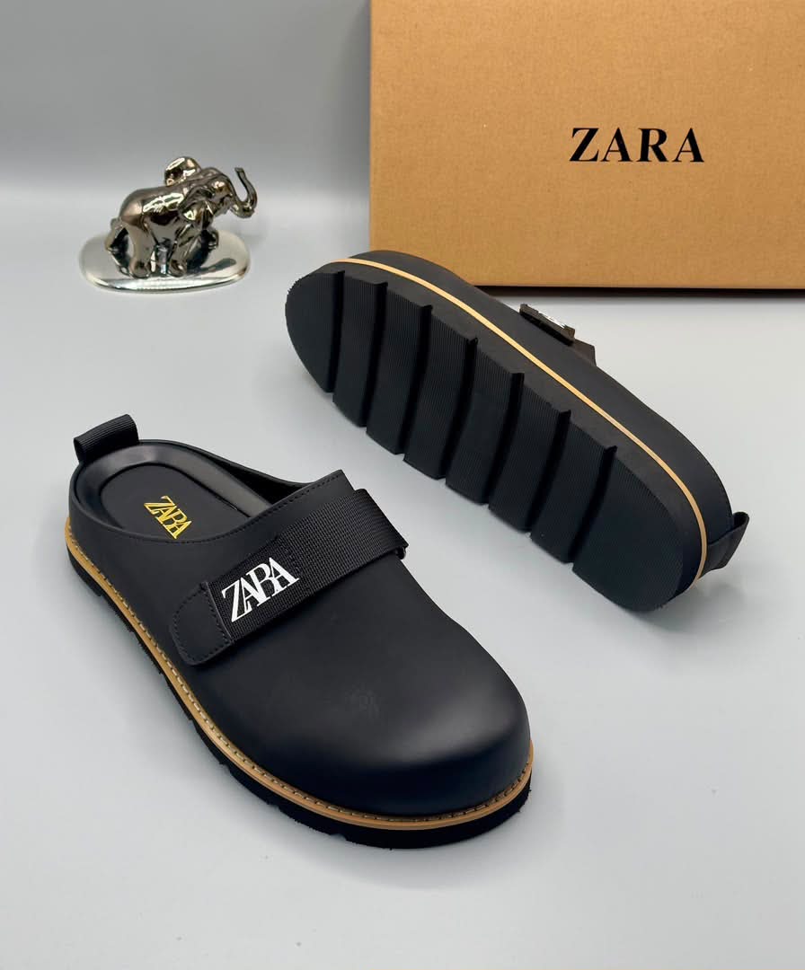 root A pair of black leather clogs with gold accents and a velcro strap, displayed on a white surface with a brown box and silver elephant figurine in the background.