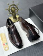 A pair of black leather monk strap shoes on a white cloth, accompanied by a shoe tree and a decorative ship's wheel in the background.