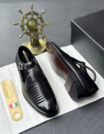 A pair of black leather monk strap shoes on a white cloth, accompanied by a shoe tree and a decorative ship's wheel in the background.