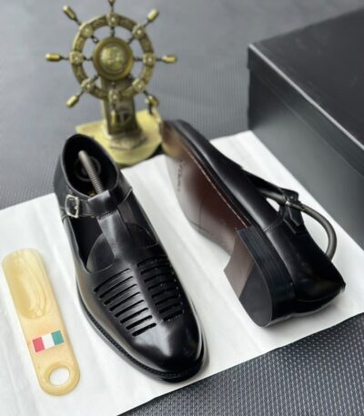 A pair of black leather monk strap shoes on a white cloth, accompanied by a shoe tree and a decorative ship's wheel in the background.